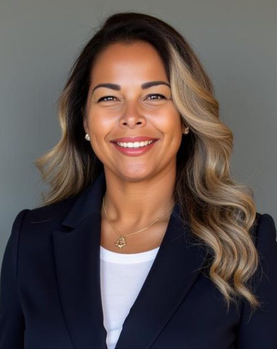 IMG_0635 A woman with long, wavy hair smiles warmly. She wears a navy blazer over a white top and a pendant necklace. The background is a plain grey.