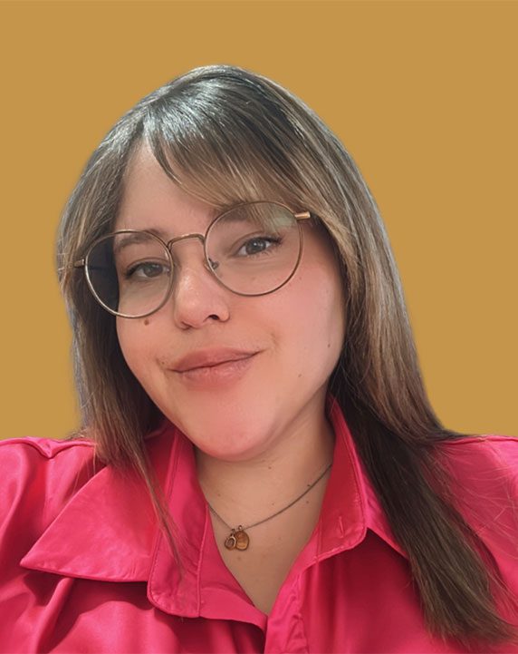 DianaLantigua A person with straight brown hair, wearing round glasses and a hot pink shirt, smiles at the camera against a plain mustard yellow background.