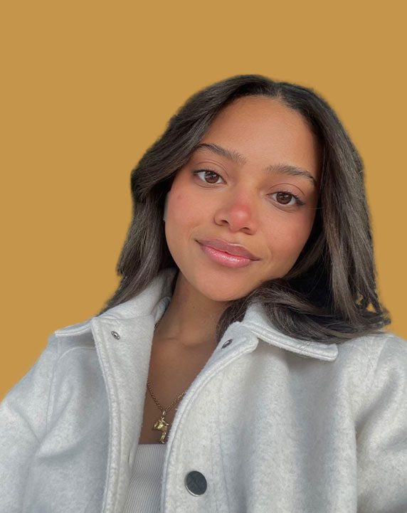 MariaFernandaSanchez A woman with long brown hair, wearing a white jacket and necklace, poses in front of a plain mustard-yellow background.